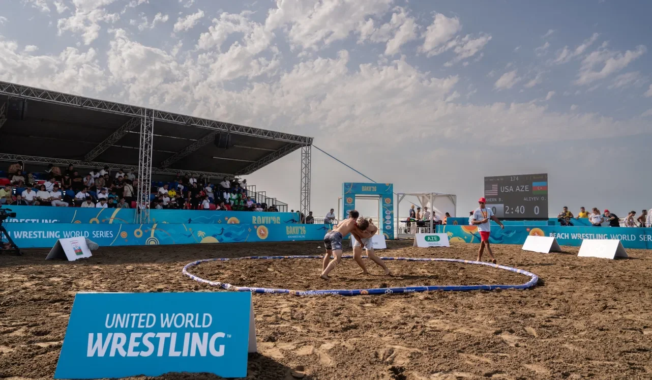 Пляжная борьба, август 2026: круг на песке, трибуна под навесом и табло United World Wrestling